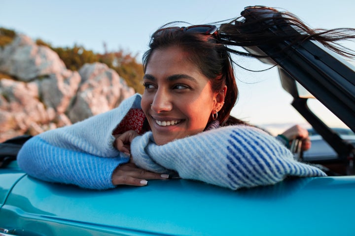 Smiling young woman day dreaming while leaning on a convertible car during a road trip.